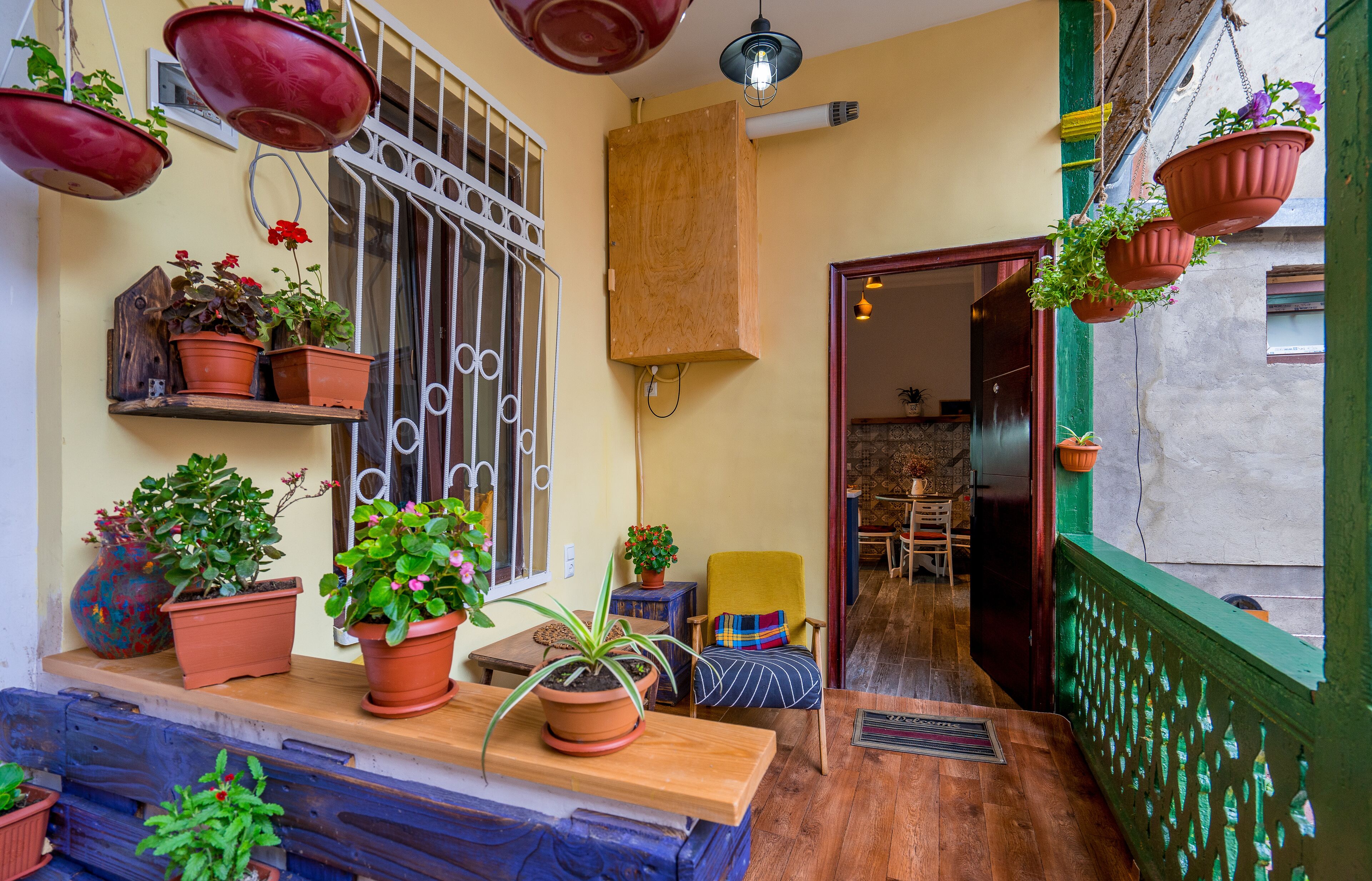 A cozy room with wooden flooring, a yellow wall, and a wooden cabinet. There are potted plants on a shelf and hanging from the ceiling.