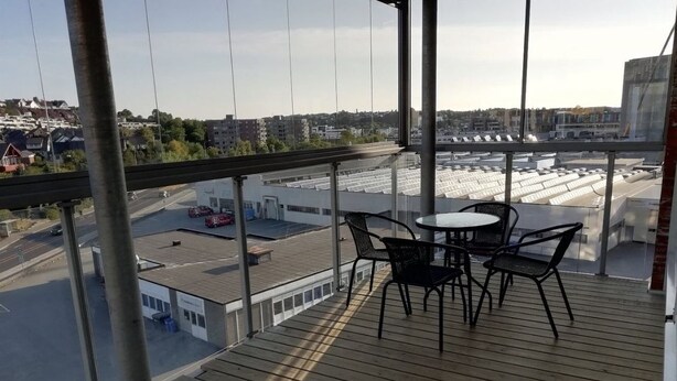 A balcony with a wooden floor, black metal chairs, and a small table. A view of a cityscape with buildings and a road is visible through the glass railing. - Gauk Apartments Sentrum 3 (Sandnes)