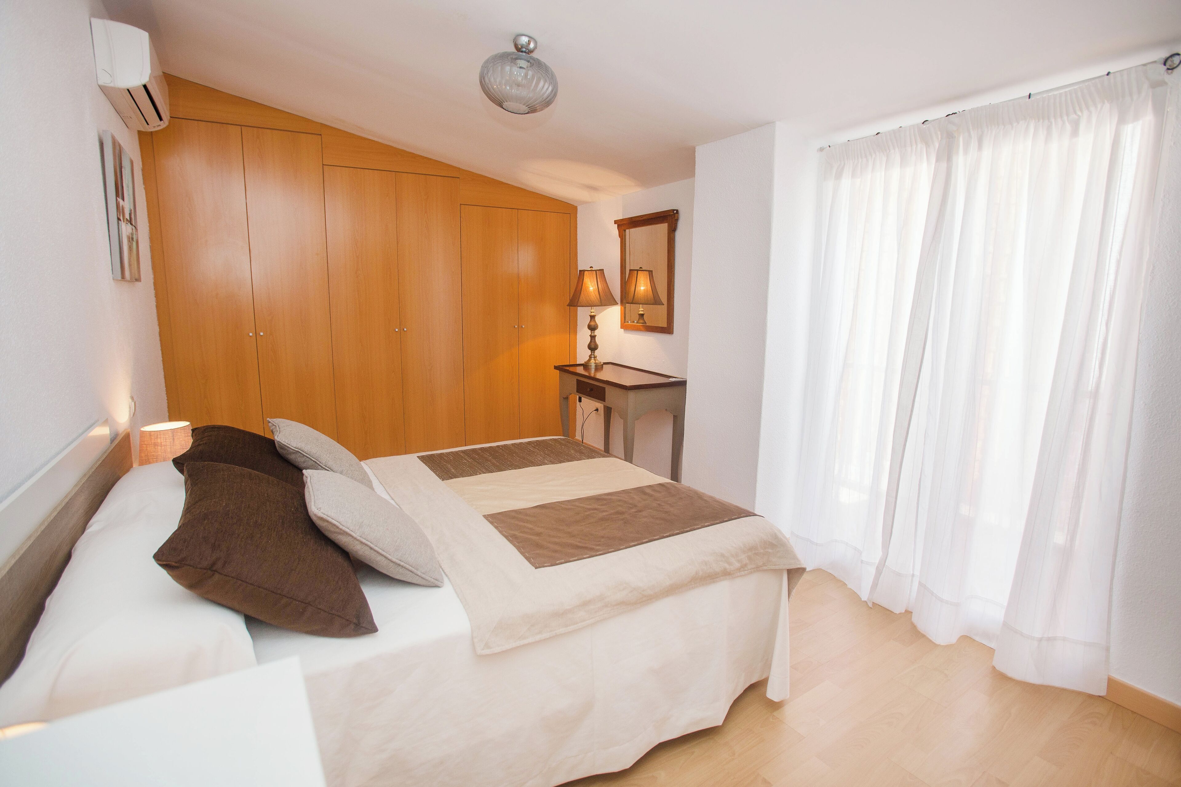 Une chambre à coucher avec un lit, une armoire en bois, un bureau avec une lampe et une fenêtre avec des rideaux.