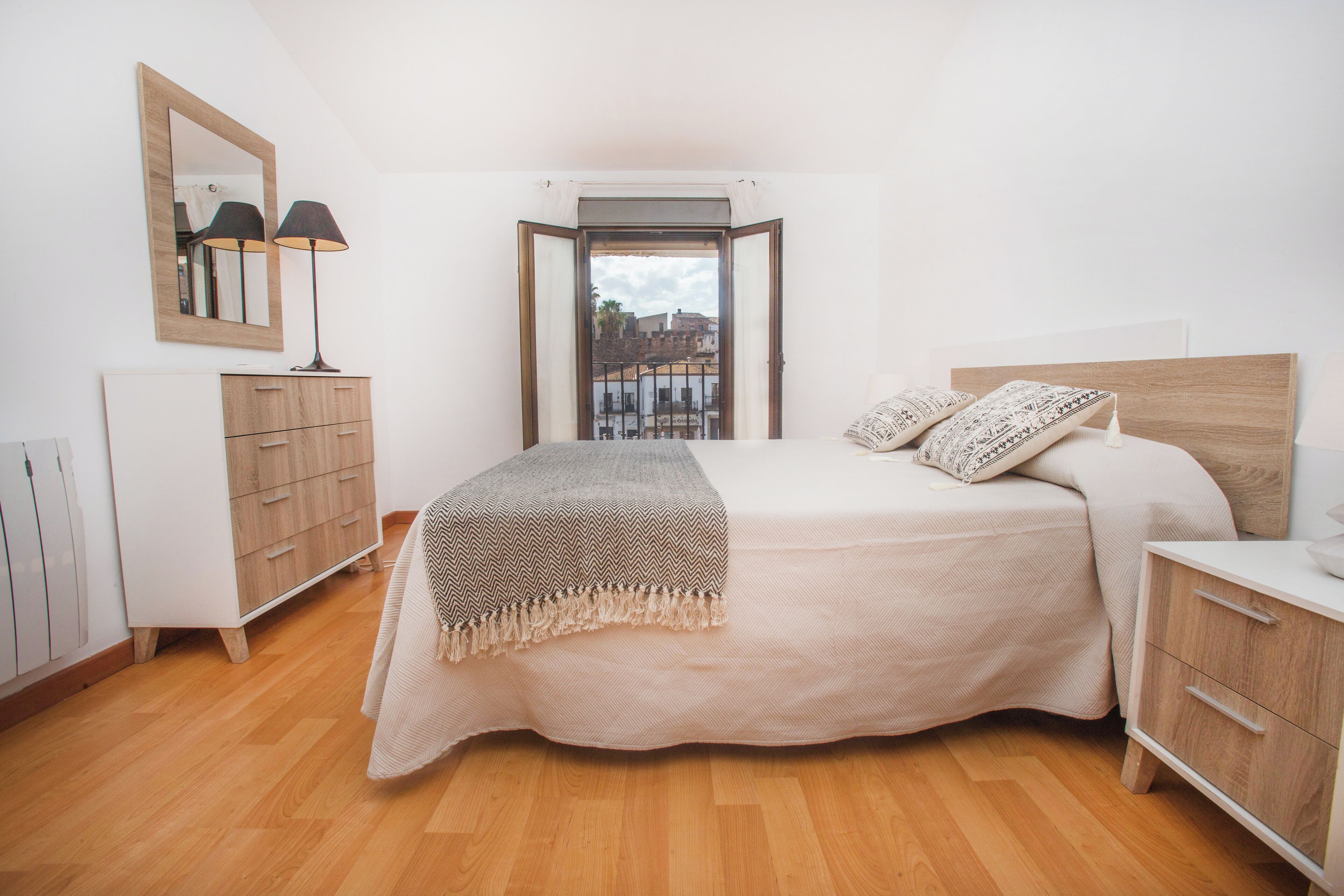 A bedroom with a bed, bedside tables, a dresser, a mirror, and a window with a view of buildings.