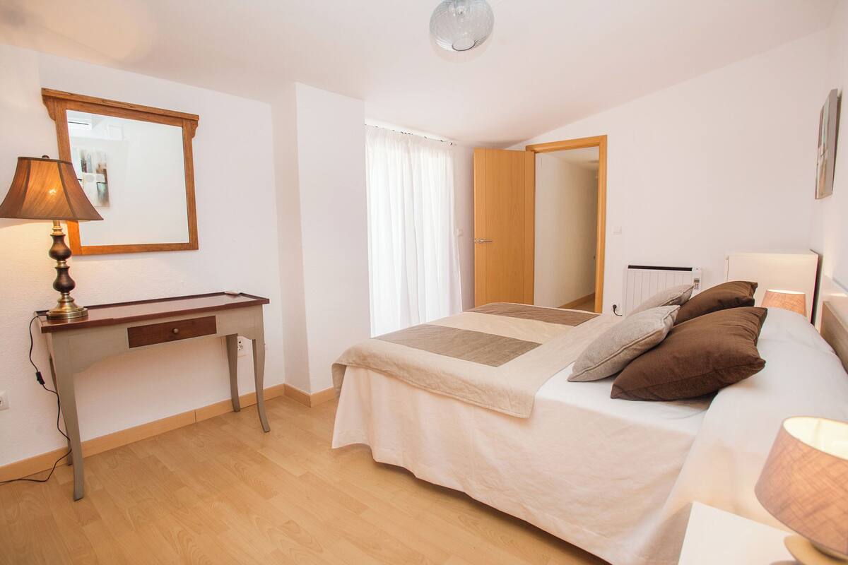 A bedroom with a bed, bedside table, lamp, and a mirror on the wall.