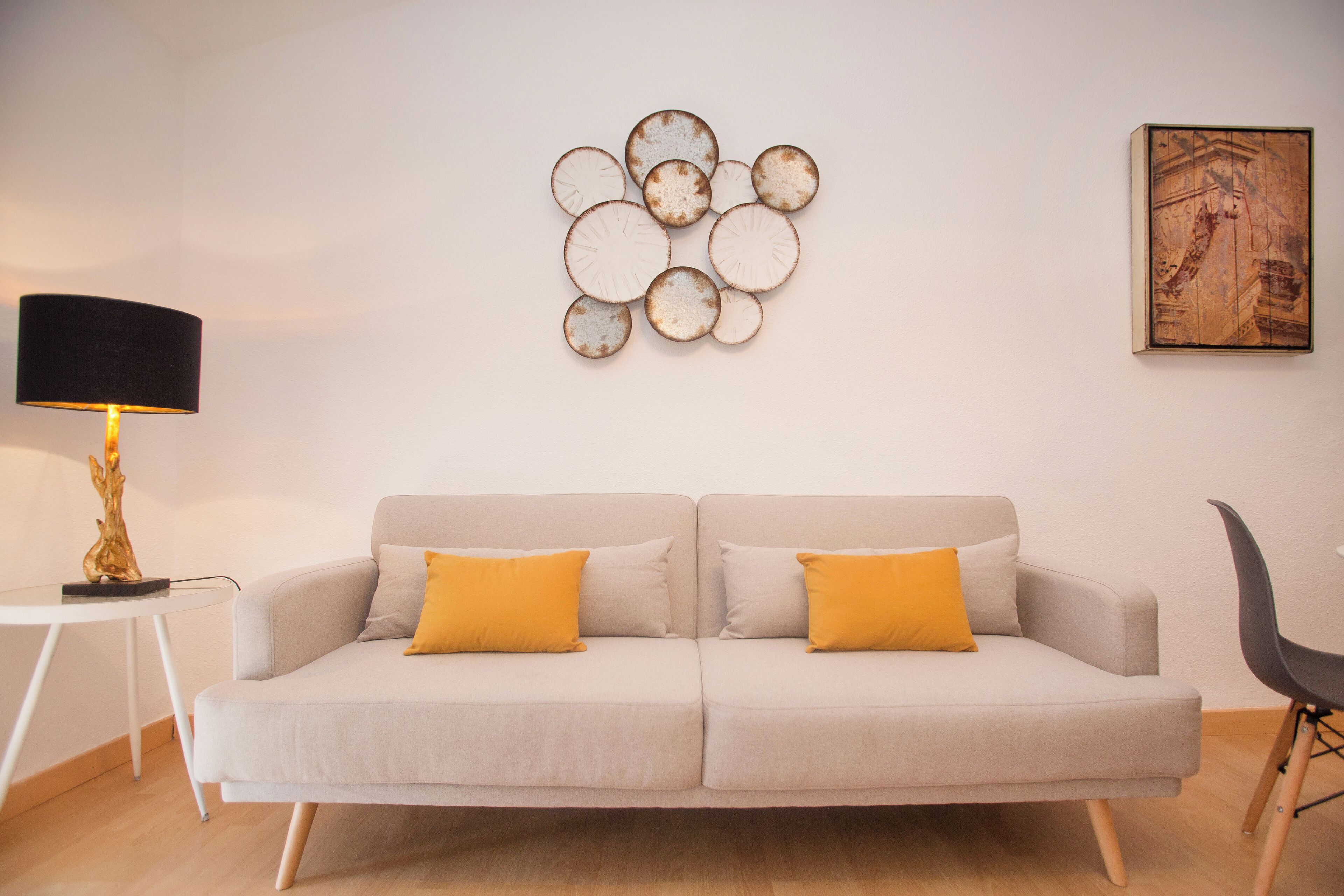 A living room with a sofa, two yellow cushions, a side table with a lamp, and a wall art piece.