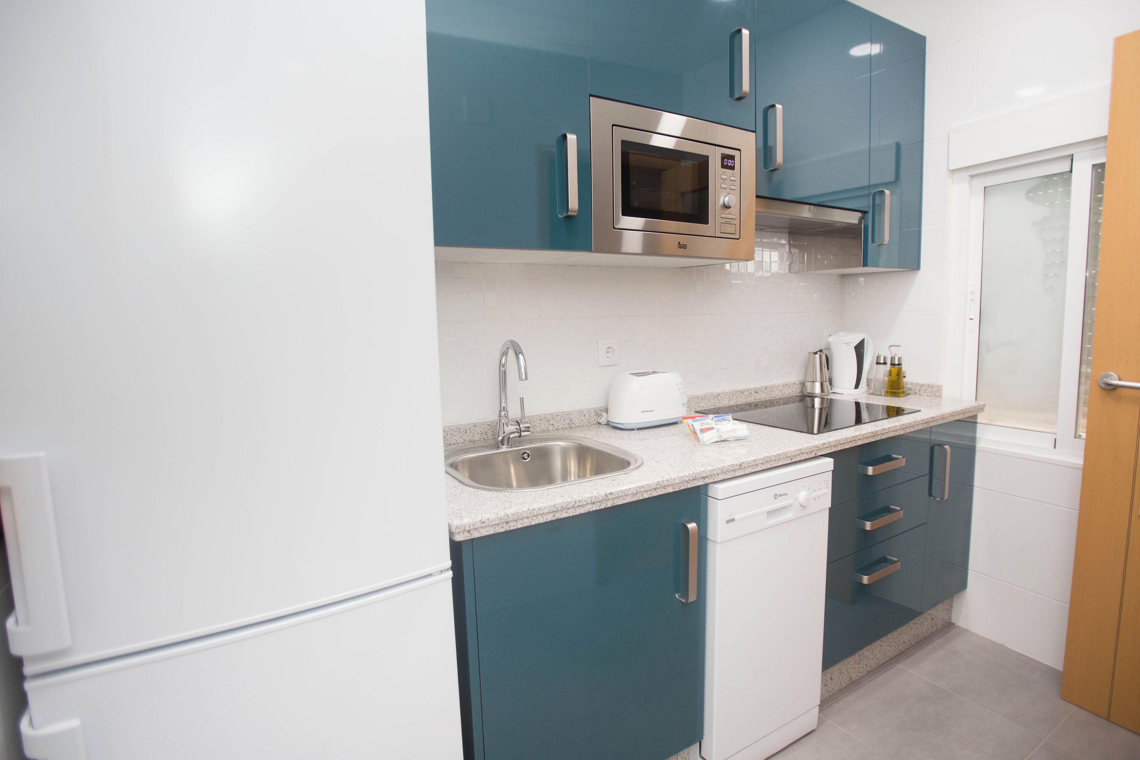 Una cocina compacta con armarios verde azulado, un microondas de acero inoxidable, un lavavajillas y un fregadero.