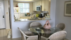 A dining area with wicker chairs and a glass table, a kitchen with white cabinets and appliances, and a dog in the hallway. - Eagle Cay Condo (Marco Island)