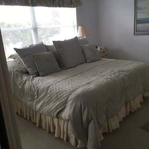 A neatly made bed with a quilted cover and ruffled edges, a bedside table with a lamp and a phone, and a window with blinds.