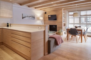 A modern kitchen with wooden cabinets, a dining area with a table and chairs, a TV mounted on the wall, and a window with curtains.
