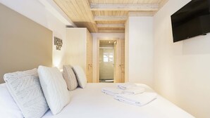 A modern hotel room with a wooden ceiling, a flat-screen TV mounted on the wall, a neatly made bed with white linens, and a glass door leading to a bathroom. - Val de Ruda Luxe 60 by FeelFree Rentals (Baqueira)