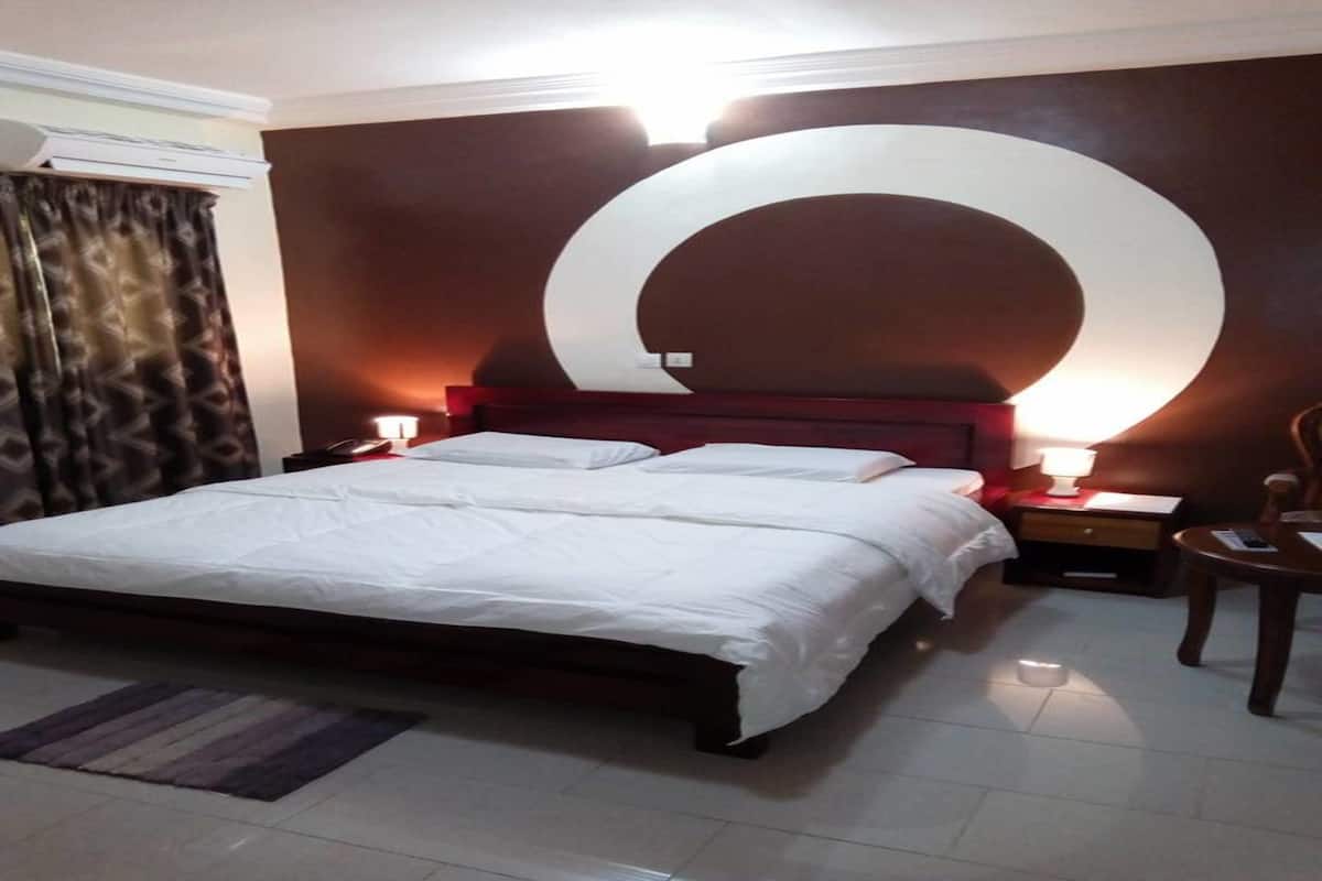 A hotel room with a large bed, two bedside tables with lamps, a wooden nightstand, a chair, and a large wall decoration.