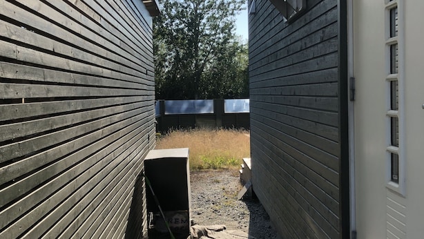 Eine schmale Holzterrasse mit Metallüberdachung, die zu einer Garage mit weißer Fassade und einem Fenster führt.