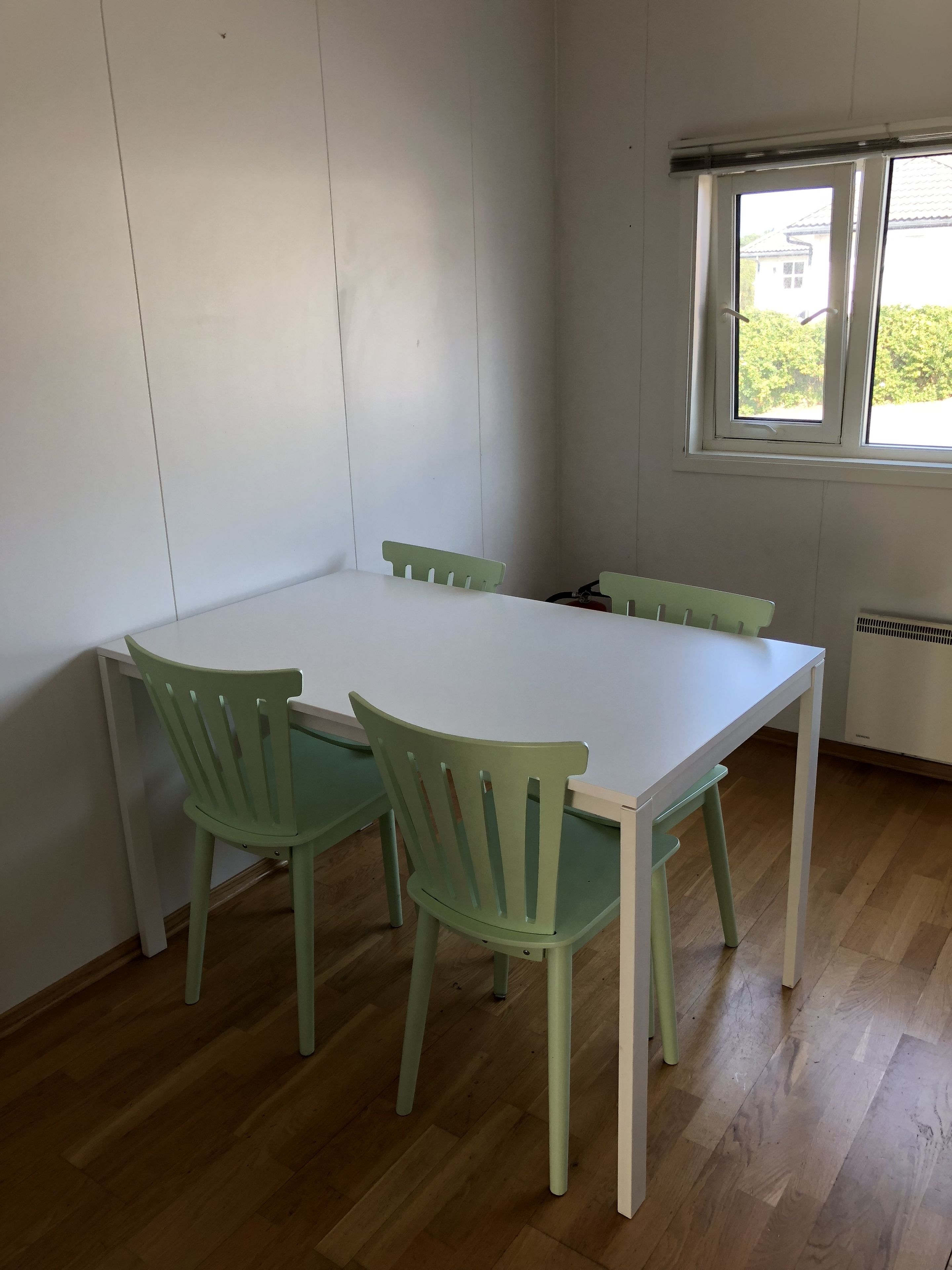 A room with a white table and four green chairs, a window with a view of greenery, and a radiator on the wall.