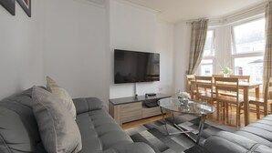 A modern living room with a grey sofa, a glass coffee table, a flat-screen TV, a wooden dining table with chairs, and a view of buildings through large windows. - Fortified Home - Near Rochester Castle (Rochester)