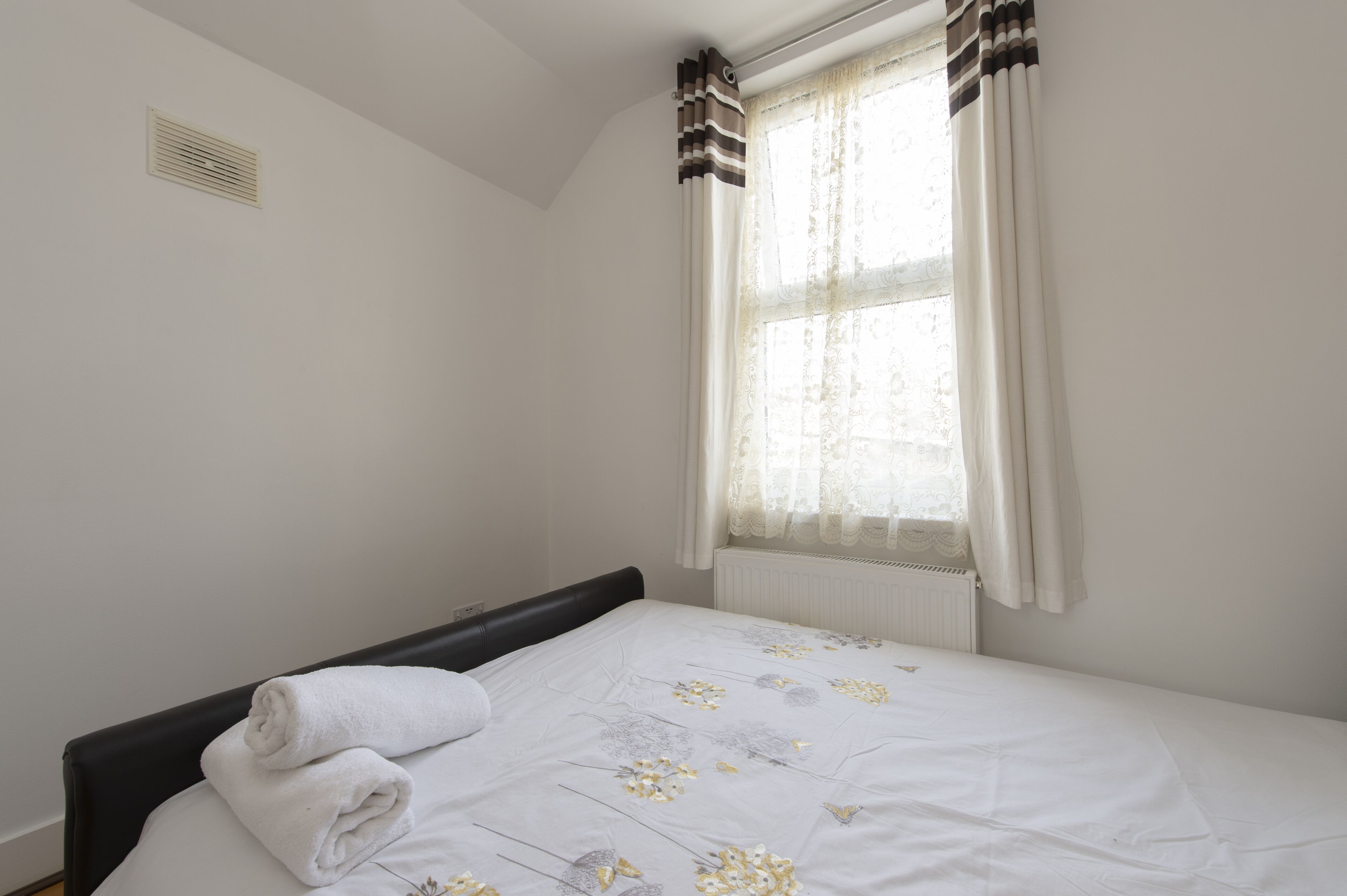 A bedroom with a bed, a window with curtains, a radiator, and a wall vent.