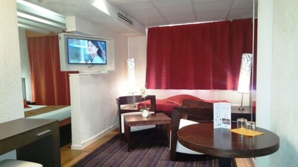 Una habitación de hotel con televisión montada en la pared, una cama con cortinas rojas, un escritorio de madera, una mesa redonda con un menú y una pequeña planta en el escritorio.