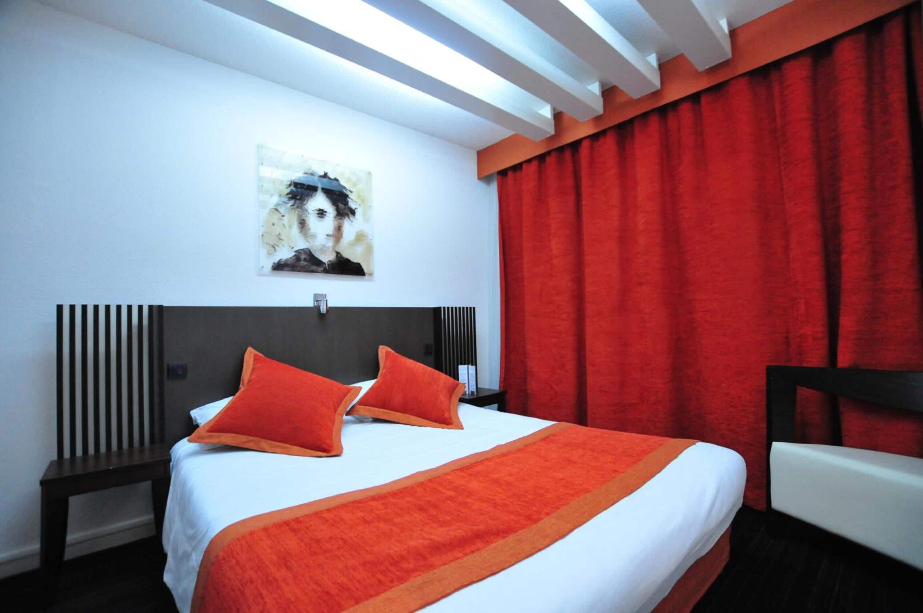A hotel room with a bed, a nightstand, a chair, a red curtain, and a painting on the wall.