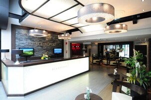 Uma área de recepção de hotel moderna, com balcão de recepção, televisão e sala de estar com assentos e plantas.