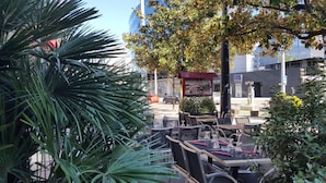 An outdoor café with tables and chairs, surrounded by greenery and a street view. - Hôtel Europole (Grenoble)