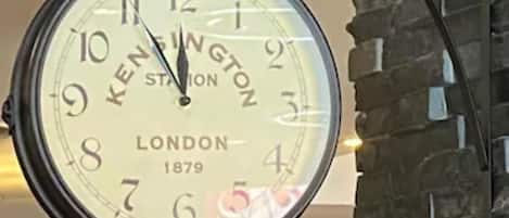 Un reloj grande sujeto a la pared, con la marca 'KENINGTON' y el año '1879' visibles, lo que denota su estilo vintage.