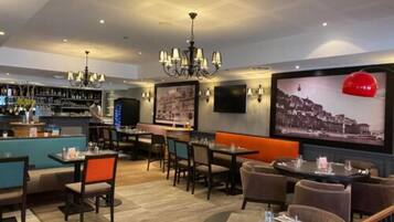 A restaurant with a bar area, multiple seating options including booths and tables, a chandelier, and a large wall-mounted TV displaying a cityscape.