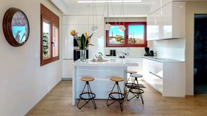 A modern kitchen with a central island, bar stools, and a view of the outdoors through a window. - Ocean Villas Complex by Ocean Residences (Malevizi)