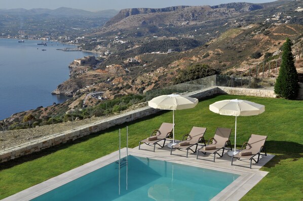 Una piscina con sillones de descanso y sombrillas, con vistas a un paisaje costero con colinas y una masa de agua.