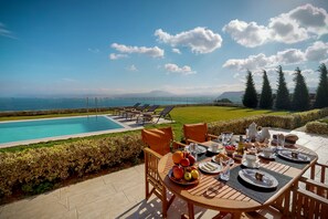 Una mesa de comedor dispuesta con platos, cubiertos y un frutero, con vista a una piscina y un paisaje escénico del mar y las montañas.