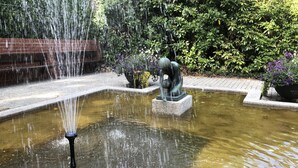 A fountain with water jets in a garden area, surrounded by greenery and a statue. - Gauk Apartments Sentrum 4 (Sandnes)