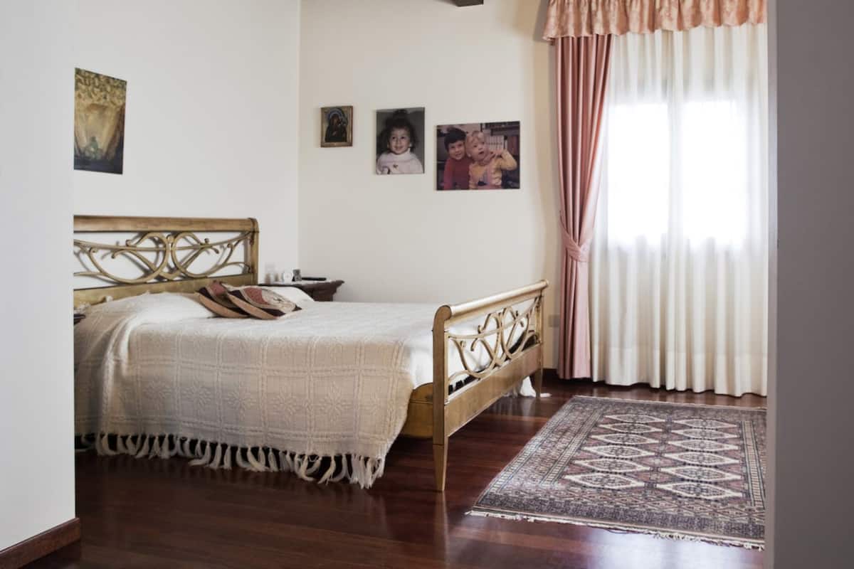 A bedroom with a wooden bed frame, a patterned bedspread, a rug, and a window with curtains.