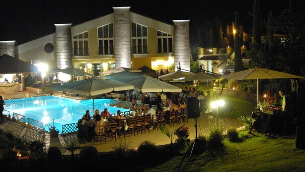 A nighttime outdoor dining area with a pool, umbrellas, and a building with large windows and columns. - Assia Spa (Fucecchio)