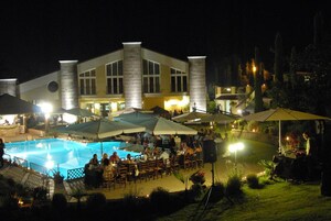 Un área de comedor al aire libre de noche con piscina, sombrillas y un edificio con amplios ventanales y columnas.