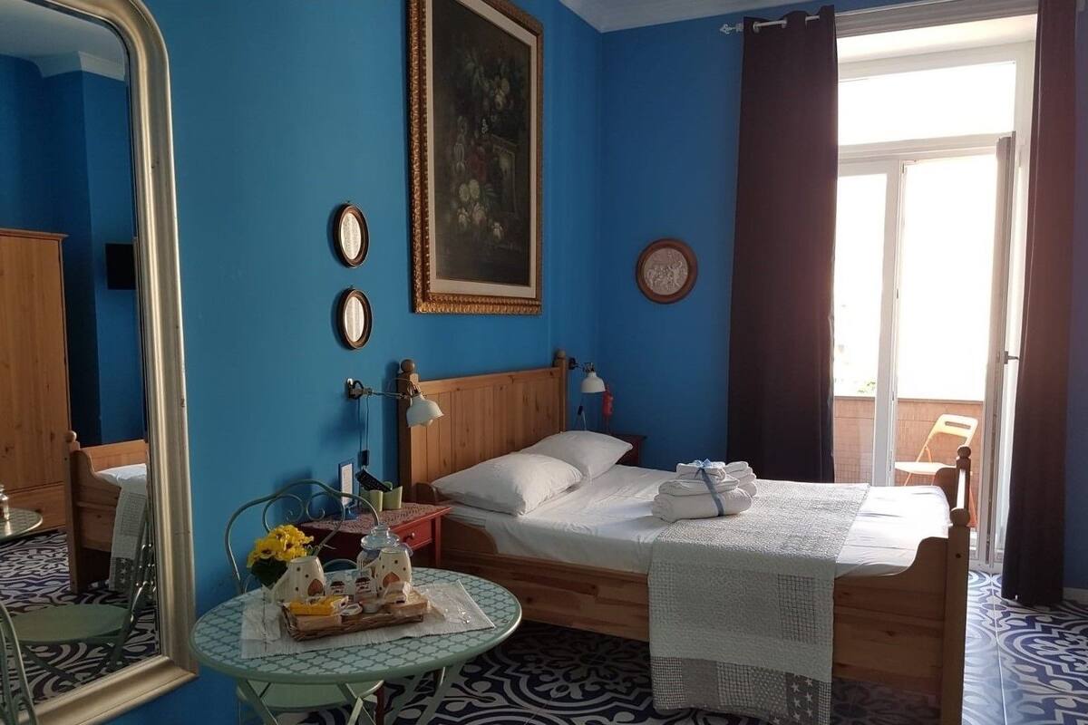 Une chambre d’hôtel avec un lit, une table ronde ornée d’un vase de fleurs, un miroir et une fenêtre avec des rideaux.
