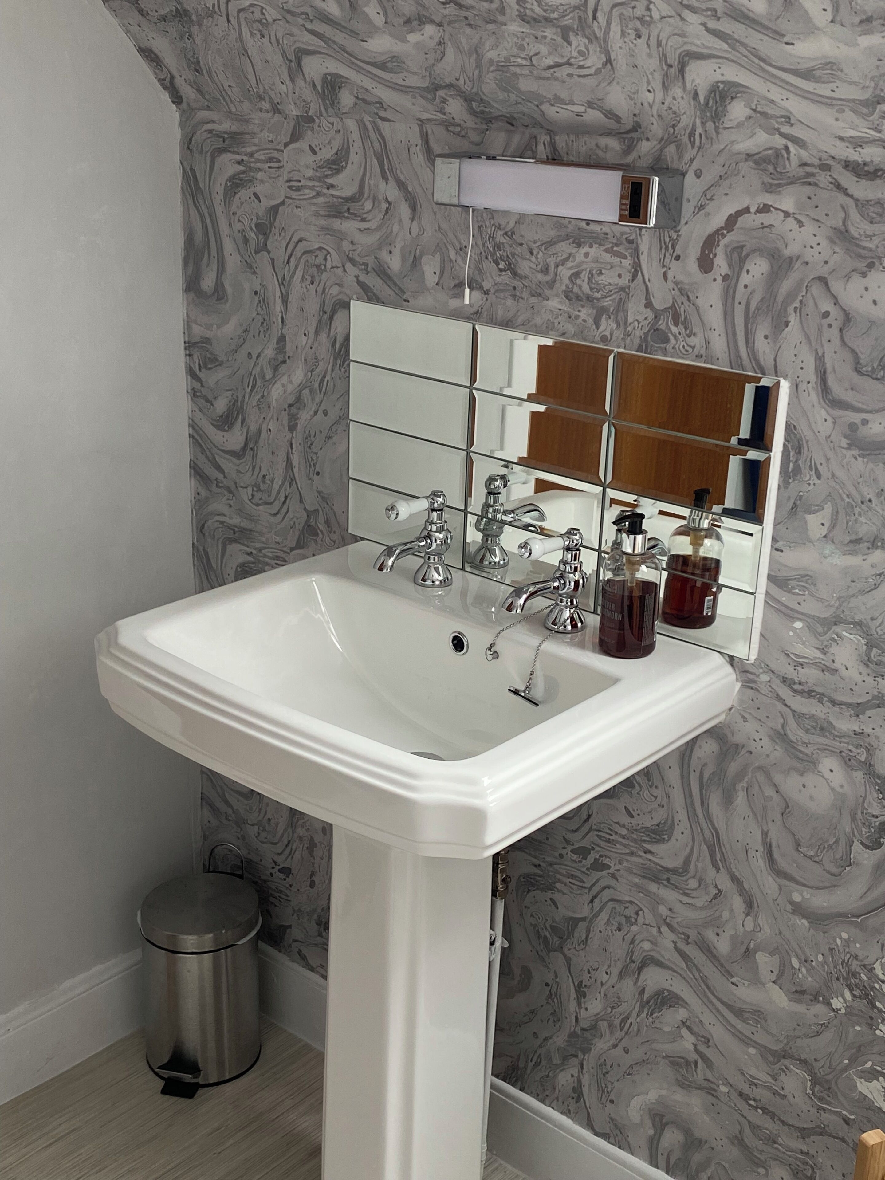 A bathroom with a white pedestal sink, a mirror with a wooden frame, and a marble-patterned wall.