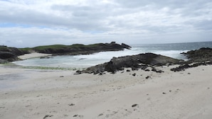 A coastal landscape with sandy beach, rocky outcrops, and grassy hills under a cloudy sky. - The Drumbeg Hotel (Drumbeg)