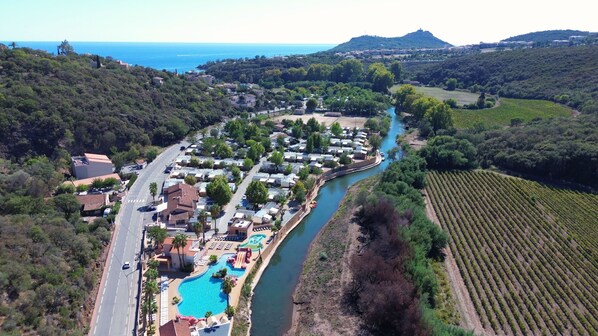 An aerial view of a coastal town with a river running through it, surrounded by vineyards and a swimming pool area. - Camping Tikayan La Vallée du Paradis (Saint-Raphael)