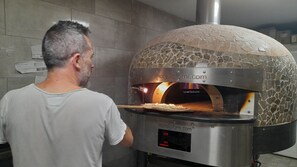 A man in a white shirt standing in front of a stone-tiled wall with a pizza oven. - Camping Tikayan La Vallée du Paradis (Saint-Raphael)