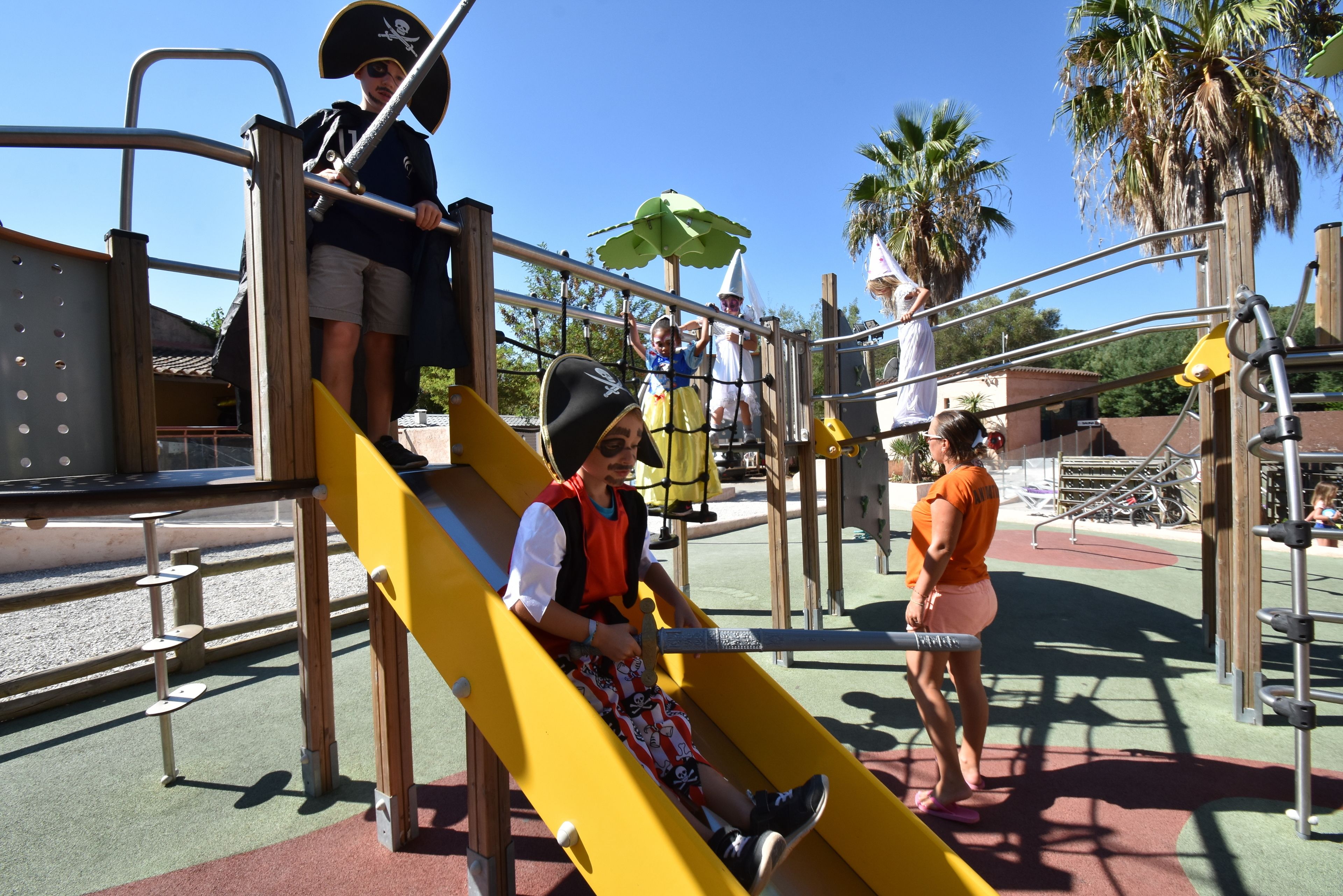 A playground with a yellow slide, climbing structures, and a palm tree in the background.