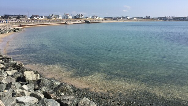 A coastal scene with a clear beach, a rocky shoreline, and a row of houses in the background. - The Haven Guest House (Holyhead)