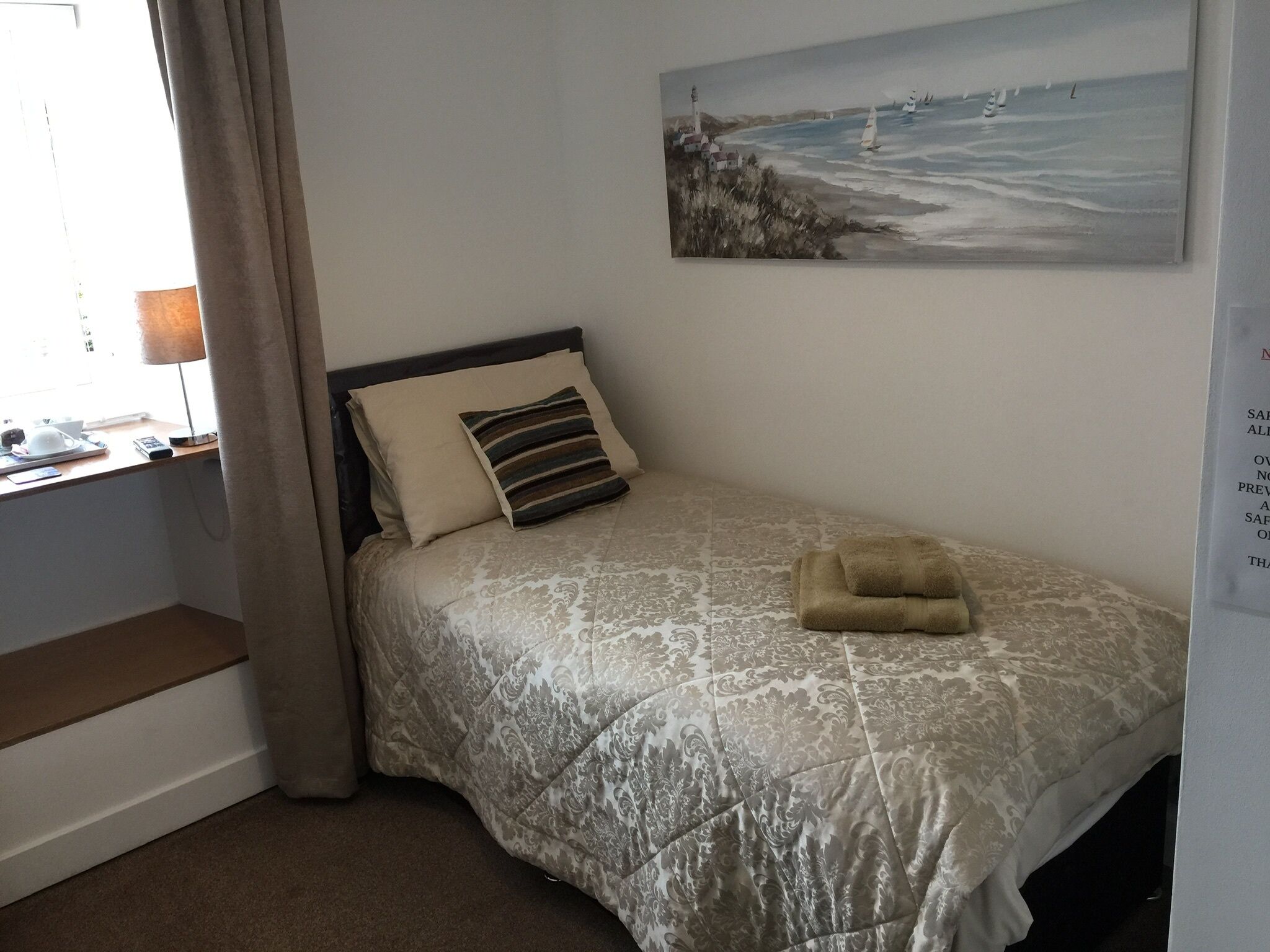 A neatly made bed with a patterned bedspread, a pillow, and a folded towel. There is a painting of a coastal scene on the wall above the bed.