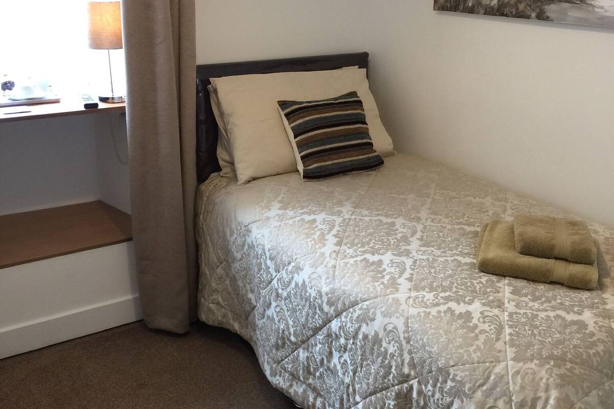 Una cama bien hecha con una colcha estampada, una almohada y una toalla doblada. En la pared hay un cuadro que representa una escena costera.