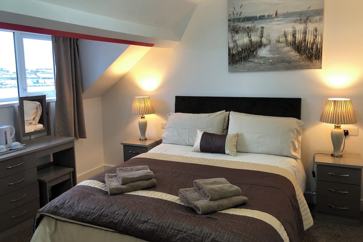 Habitación de hotel con una cama grande, dos mesitas de noche con lámparas, un escritorio con silla, una ventana con cortinas y un cuadro en la pared.