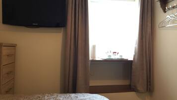 A bedroom with a bed, a flat-screen TV mounted on the wall, a window with curtains, a bedside table with a lamp, and a wooden dresser.