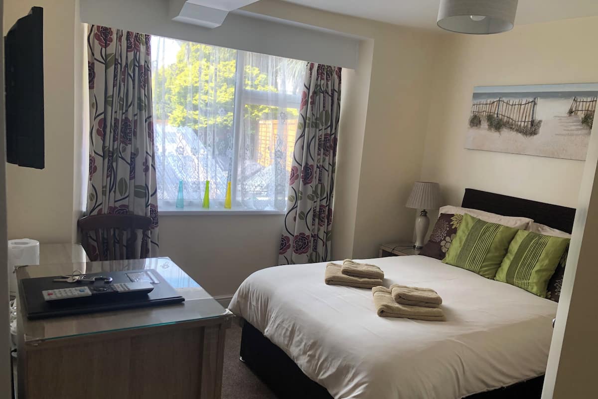 Habitación de hotel con cama, escritorio, silla, ventana con cortinas, lámpara y un cuadro en la pared.