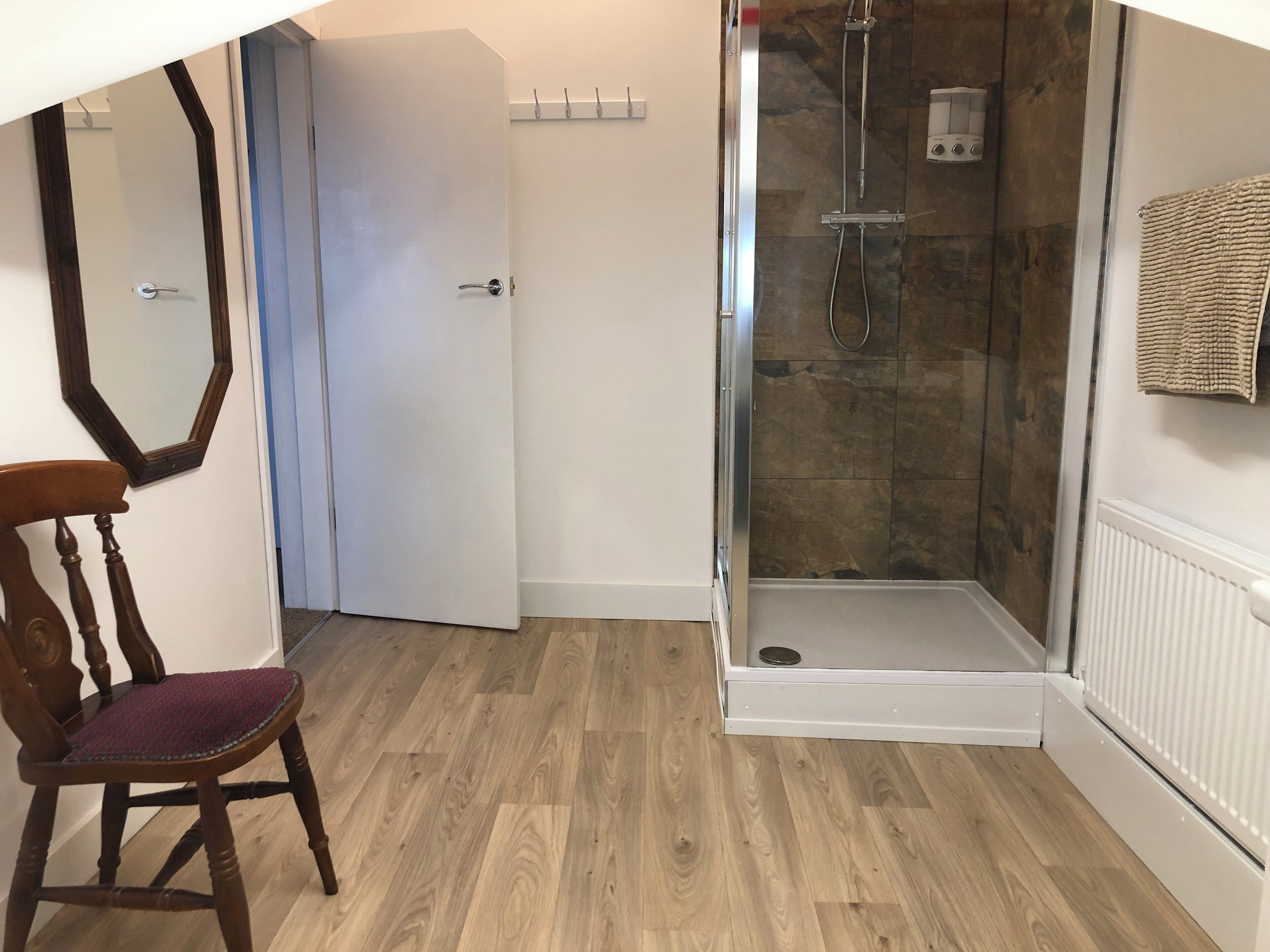 A room with a wooden floor, a white door, a shower with a glass door, and a towel hanging on the wall.