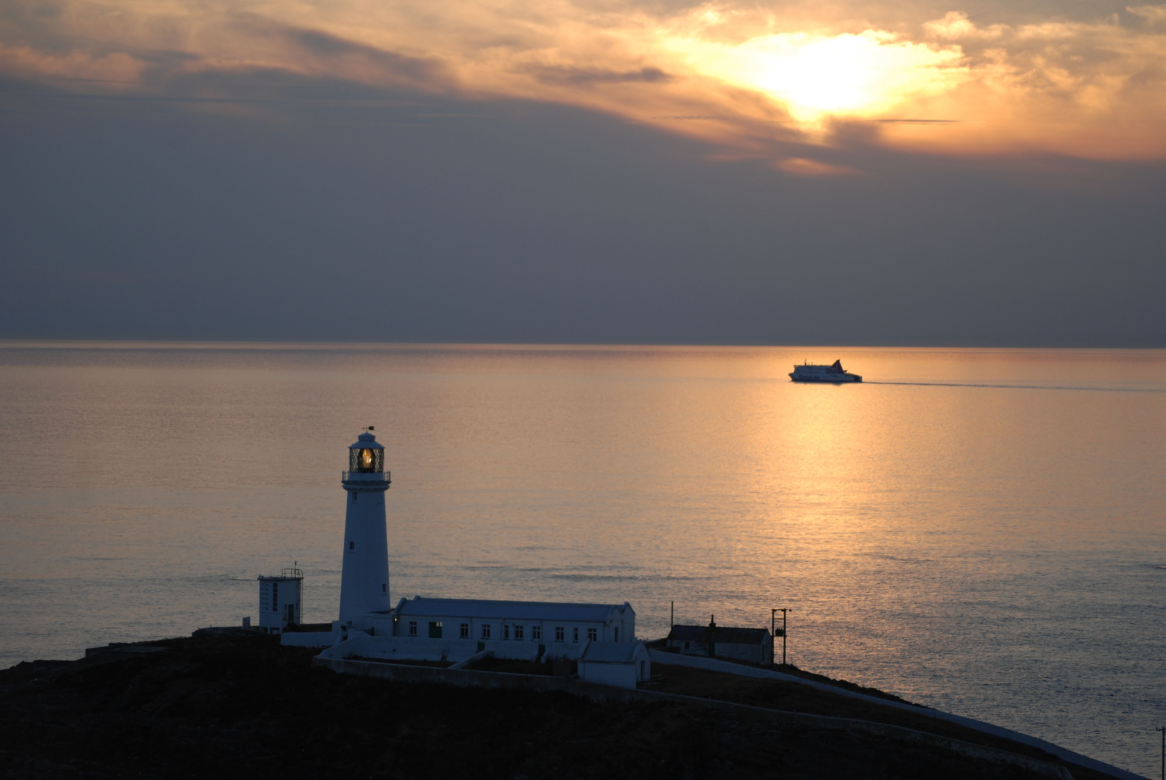 A lighthouse at sunset with calm sea and a ship in the distance.