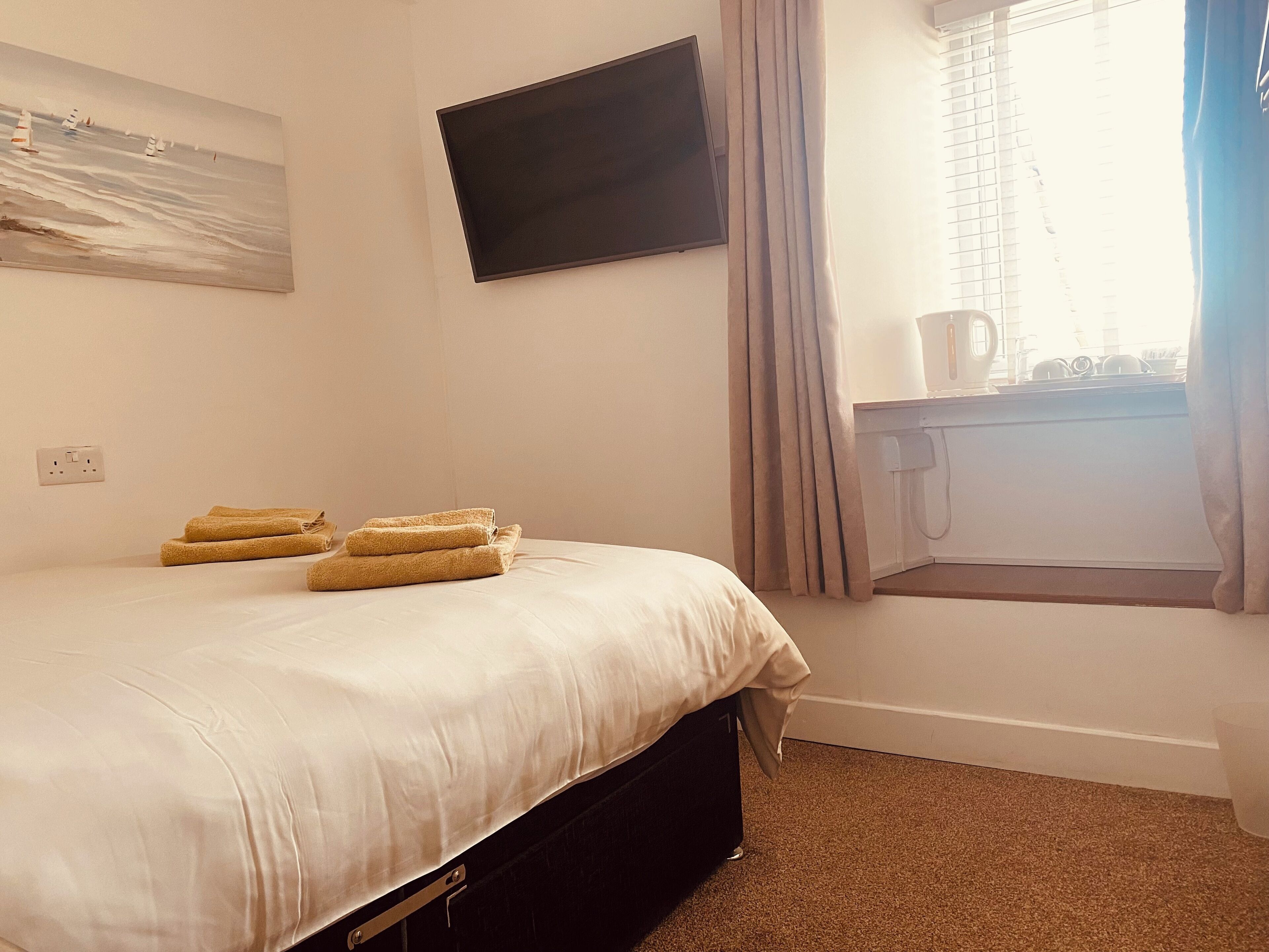 A hotel room with a bed, a flat-screen TV, a window with curtains, and a small table with a kettle.