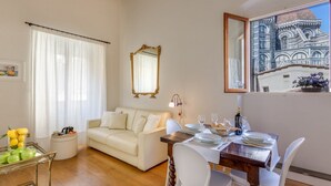 A living room with a white sofa, a dining table set for two, and a large window with a view of a building. - Benedetto Duomo (Florence)