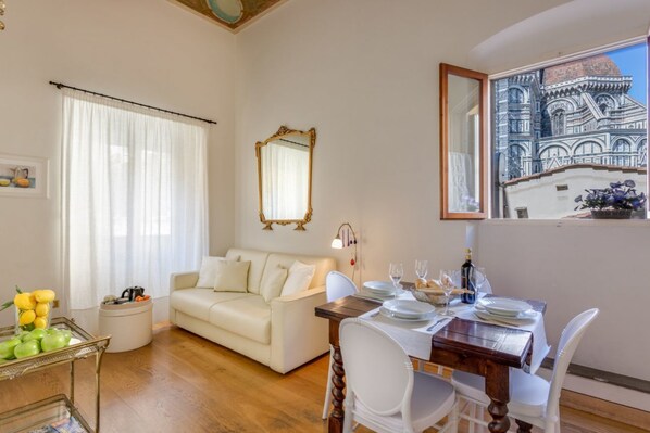 A living room with a white sofa, a dining table set for two, and a large window with a view of a building.