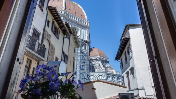 A narrow street with buildings featuring ornate facades and a prominent dome structure. - Benedetto Duomo (Florence)