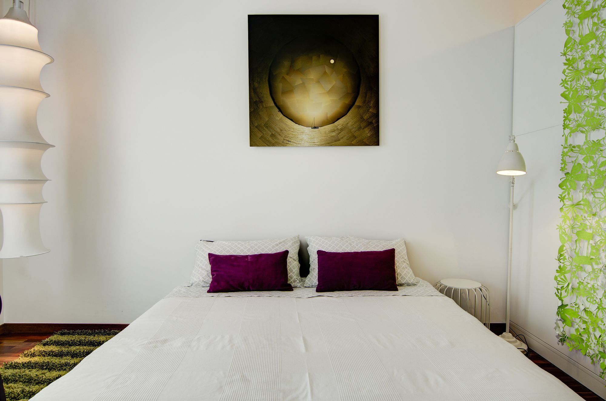 A bedroom with a bed, two pillows, a wall-mounted artwork, a floor lamp, and a green wall decoration.