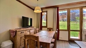 A dining area with a wooden table and chairs, a television on a wooden cabinet, and large windows with a view of buildings and greenery. - 3-room apartment, 2 bathrooms, direct access to slopes (Plagne Soleil)