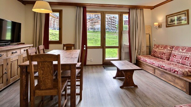 A cozy room with a wooden dining table, chairs, a sofa, a television, and large windows offering a view of buildings and greenery. - 3-room apartment, 2 bathrooms, direct access to slopes (Plagne Soleil)